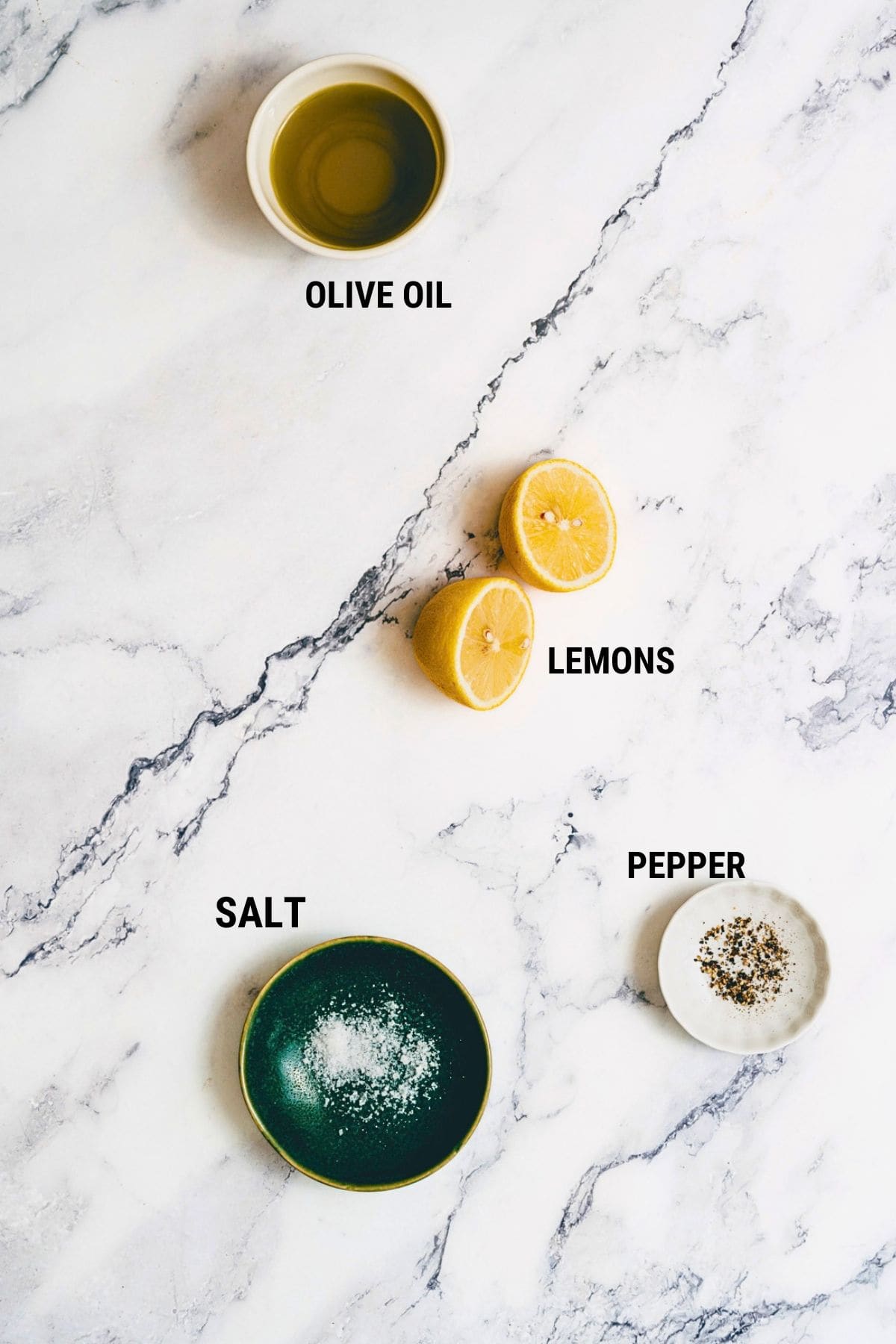A top-down view of ingredients on a marble surface: olive oil, halved lemons, salt, and pepper, each labelled with its name.