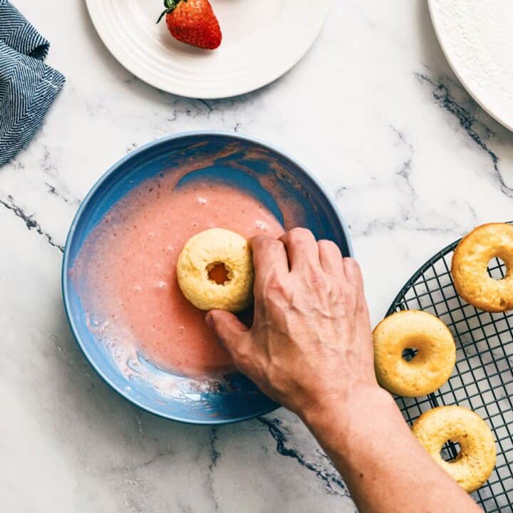 A hand dips a plain doughnut into a bowl of pink icing, next to a wire cooling rack with several other doughnuts.