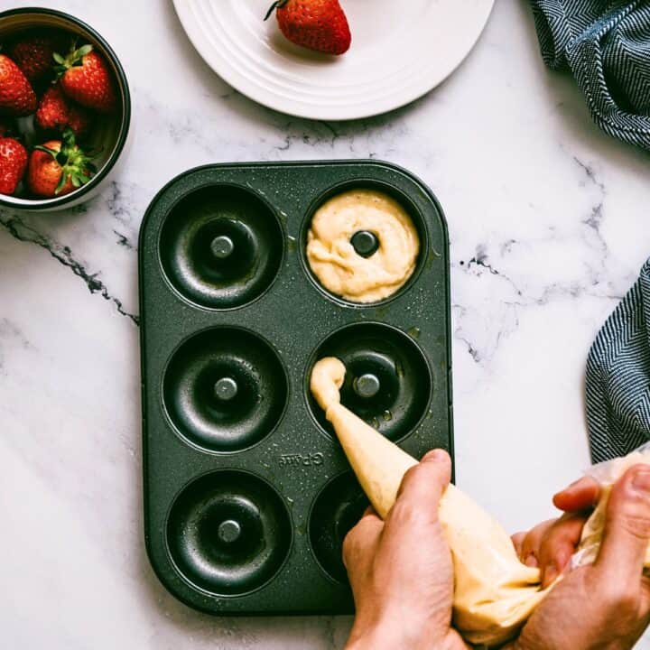 Hands using a piping bag to fill a doughnut tin with batter next to a bowl of fresh strawberries.
