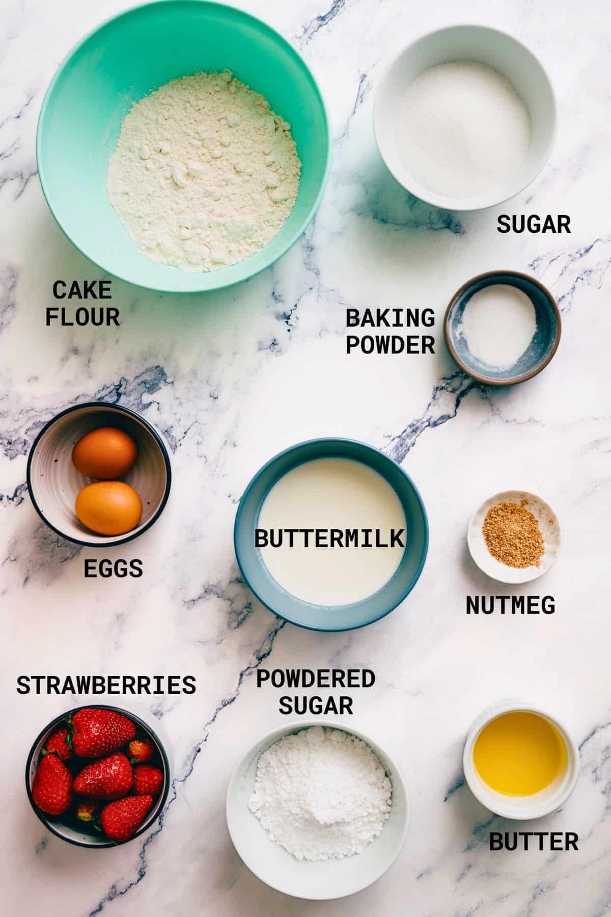 Baking ingredients for strawberry glazed doughnuts arranged on a marble surface: self-raising flour, sugar, baking powder, eggs, buttermilk, nutmeg, strawberries, icing sugar, and melted butter, each labelled with its name.