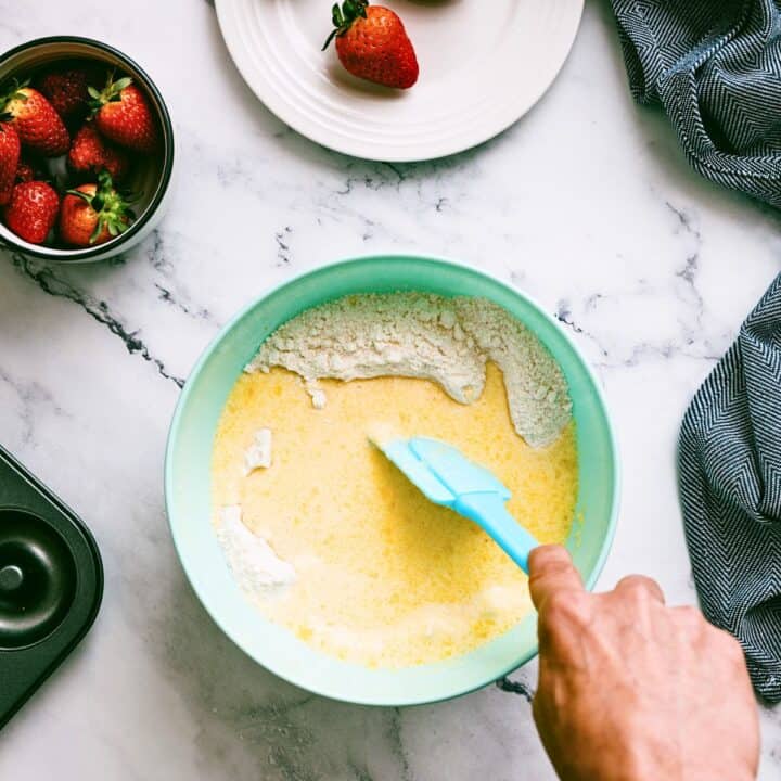 A hand uses a blue spatula to mix flour into a yellow liquid batter in a light blue bowl, with strawberries and a baking tin nearby.